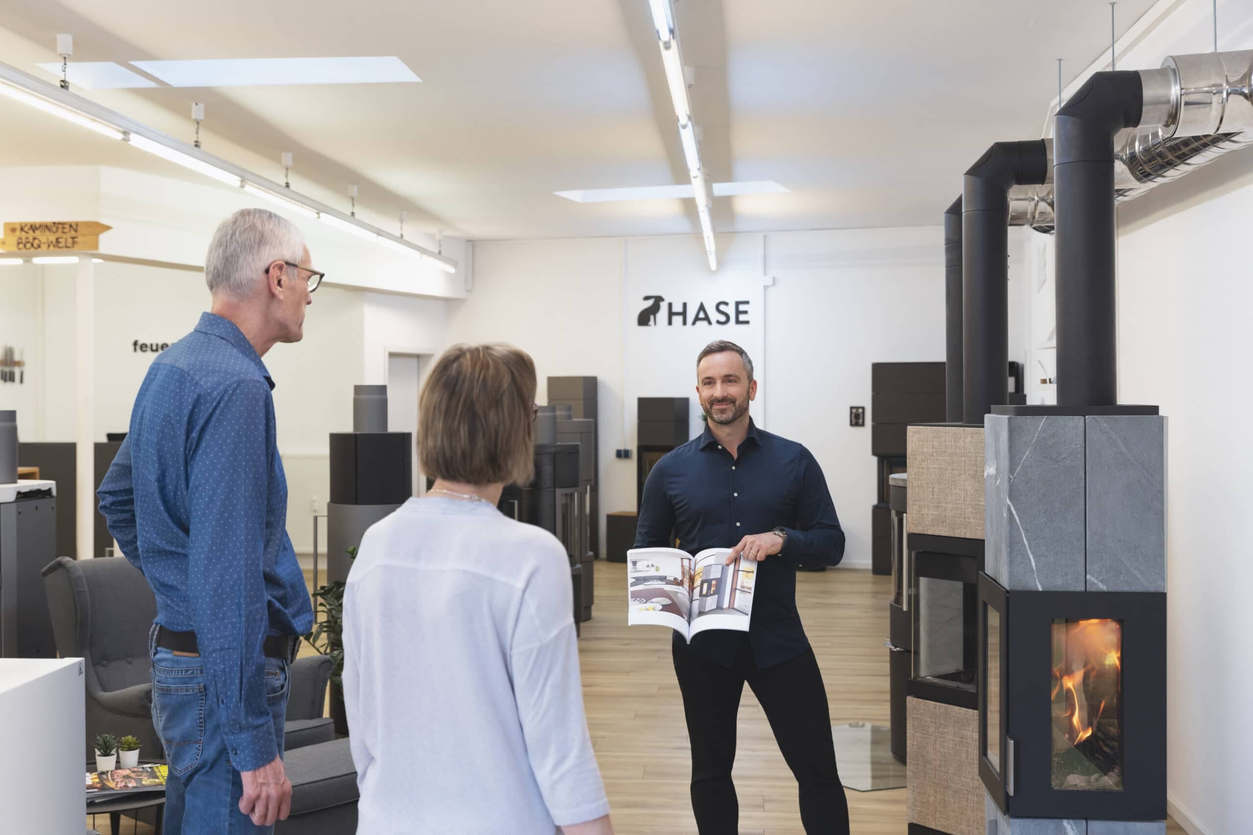 Ausstellungsraum mit Kaminöfen: Verkäufer zeigt Prospekt zwei Kunden, im Hintergrund mehrere Kaminöfen ausgestellt, einer