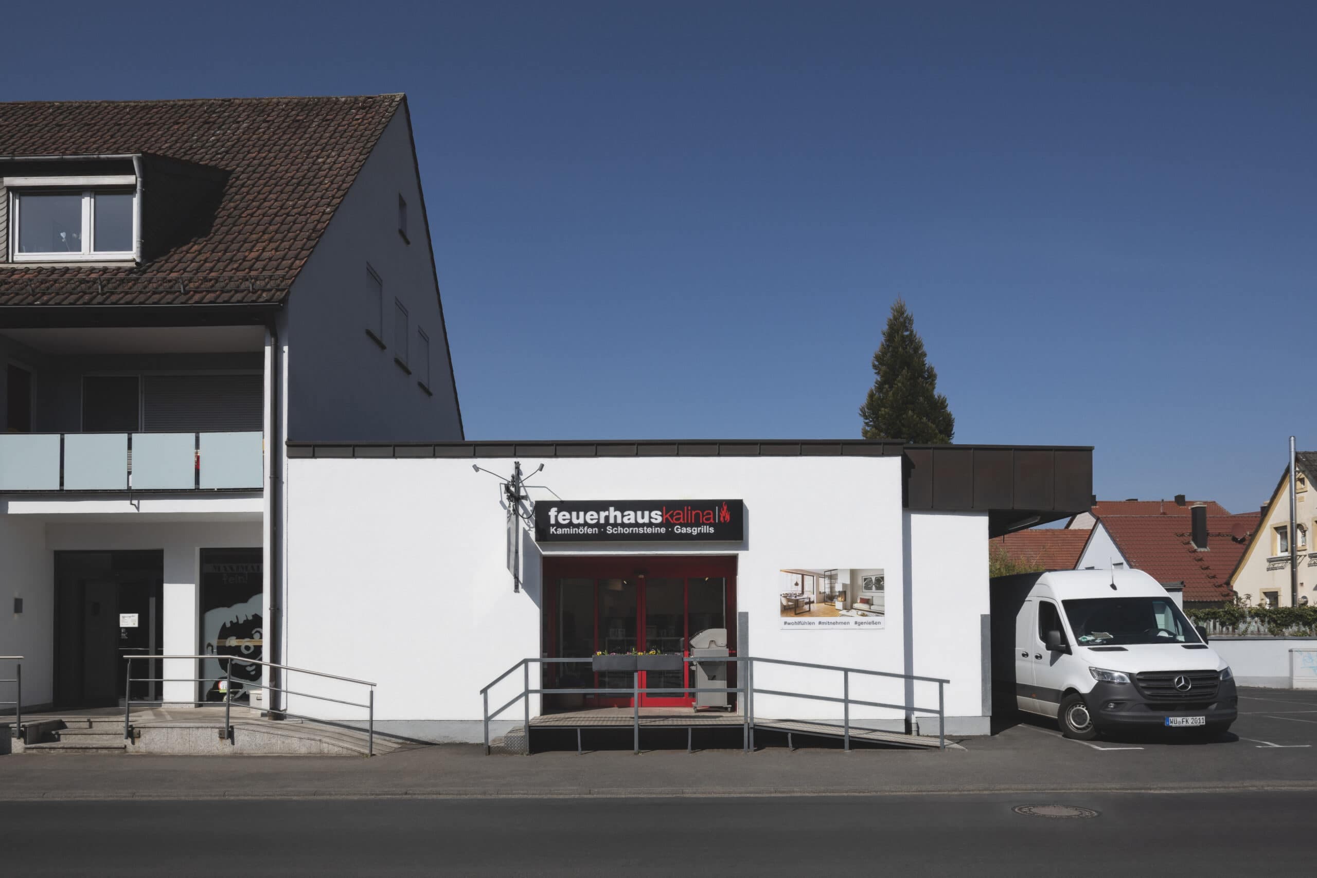 Ausstellungsgebäude von Feuerhaus Kalina in Rimpar mit weißer Fassade, rote Eingangstür und Schaufenster, daneben moderner