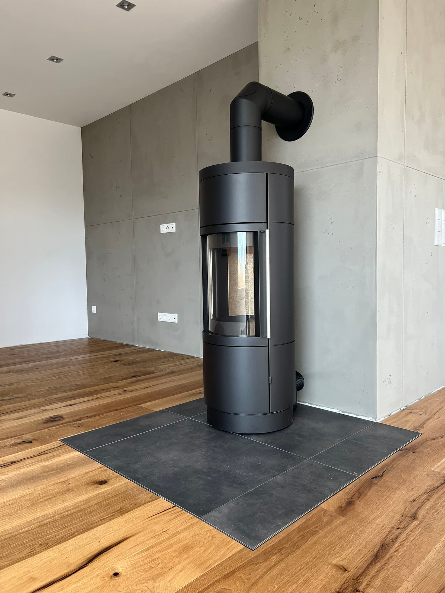 Schwarzer Kaminofen mit Glastür und Speichersteinen in modernem Wohnraum mit Betonwand und Holzboden