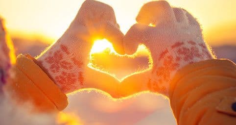 Hände in Winterhandschuhen formen ein Herz vor der Sonne bei Sonnenaufgang