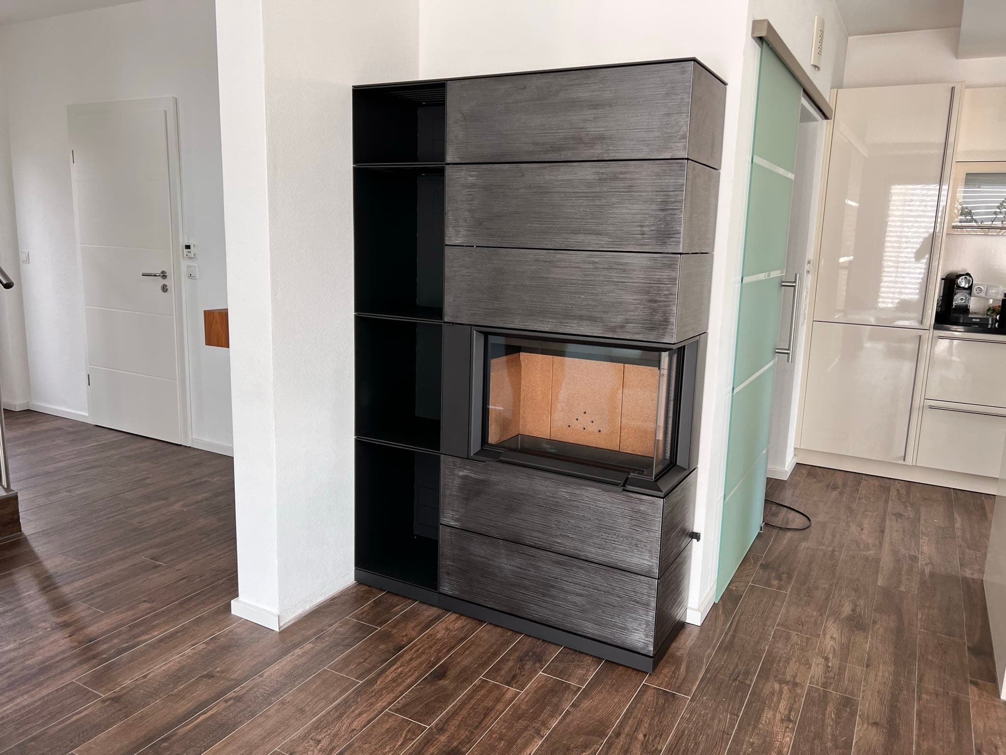 Speicherofen mit dunkelgrauer Keramik-Verkleidung und integriertem Kaminofen, Regalsystem mit offenen Fächern, in modernem