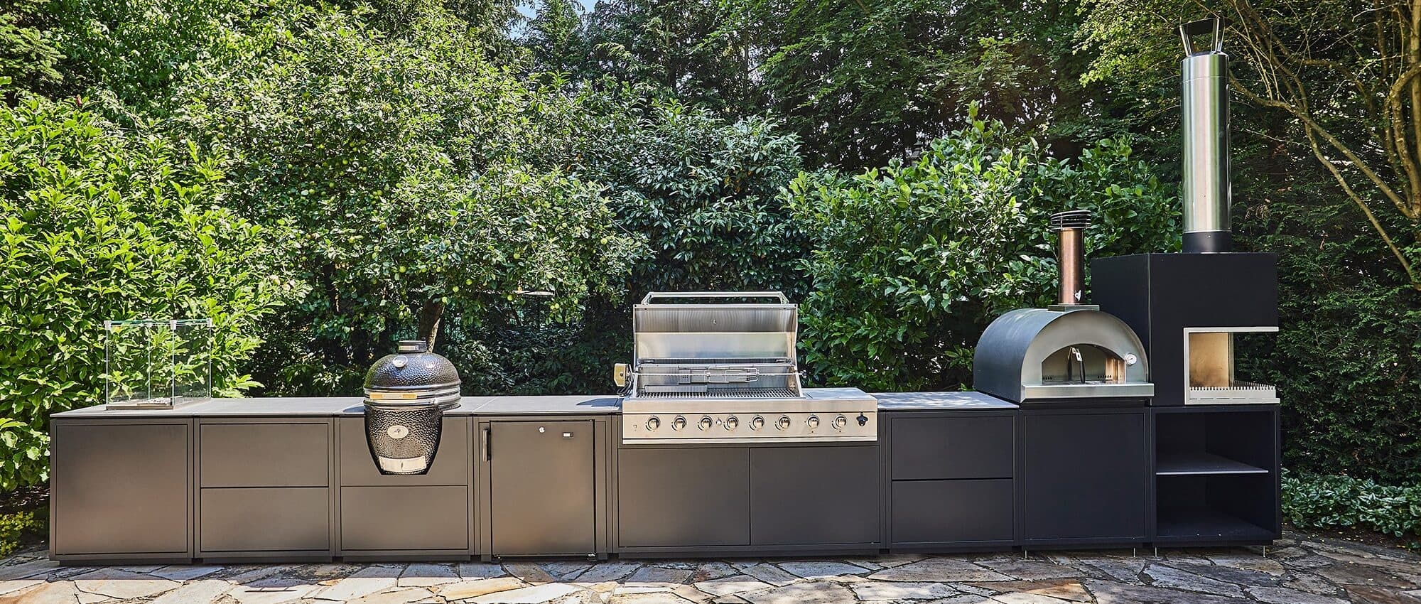 Outdoor-Küche mit Gaskochfeld, Pizzaofen, Grill und Lagerschränken vor Heckenkulisse.