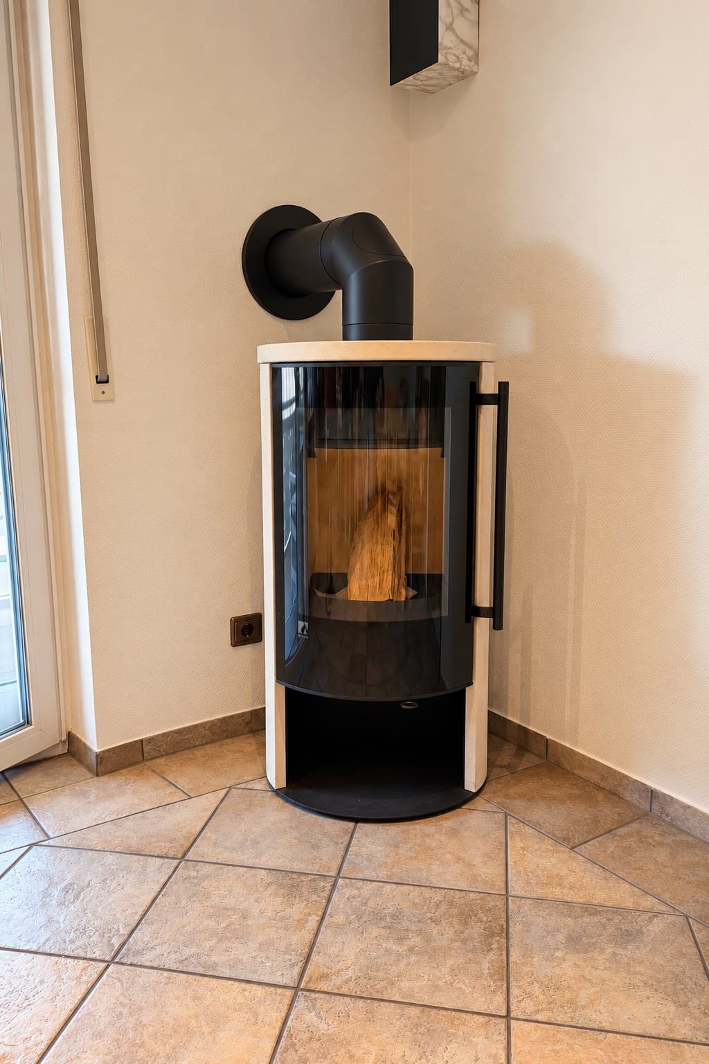 Drooff Aprica 2 Trend in einer Raumecke: runder schwarzer Korpus mit gebogener Scheibe, seitlich und oben die helle Verkleidung Premium White, darunter ein offenes Fach, das Rauchrohr führt nach hinten in die Wand.