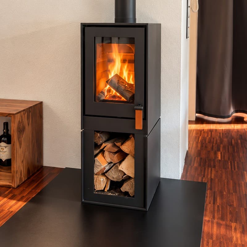 Barbas BOX 20/35 in schwarzem Stahl, freistehend mit offenem Holzfach voll Brennholz, brennendes Feuer hinter der hochkant stehenden Scheibe, brauner Lederriemen am Türgriff, aufgestellt auf einer schwarzen Stahlplatte auf Parkett.