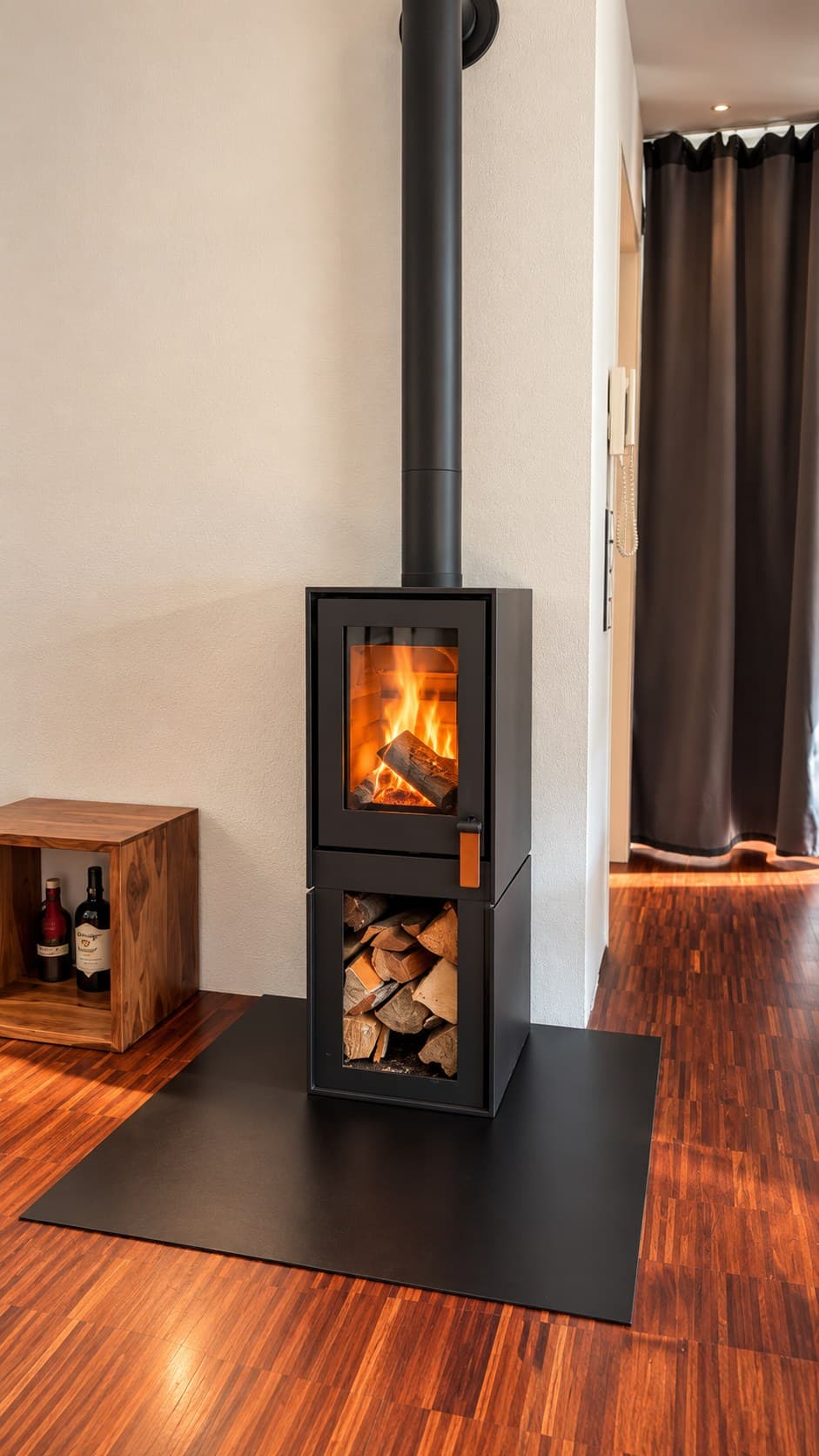 Barbas BOX 20/45 in schwarzem Stahl mit brennendem Holzfeuer hinter der hohen schmalen Scheibe, darunter das offene Holzfach voll Brennholz. Der Türgriff ist ein schwarzer Bügel mit braunem Lederriemen. Der Ofen steht auf einer schwarzen Stahlplatte, die auf dem rotbraunen Parkett aufliegt, das Rauchrohr führt nach oben und im Bogen in die Wand.