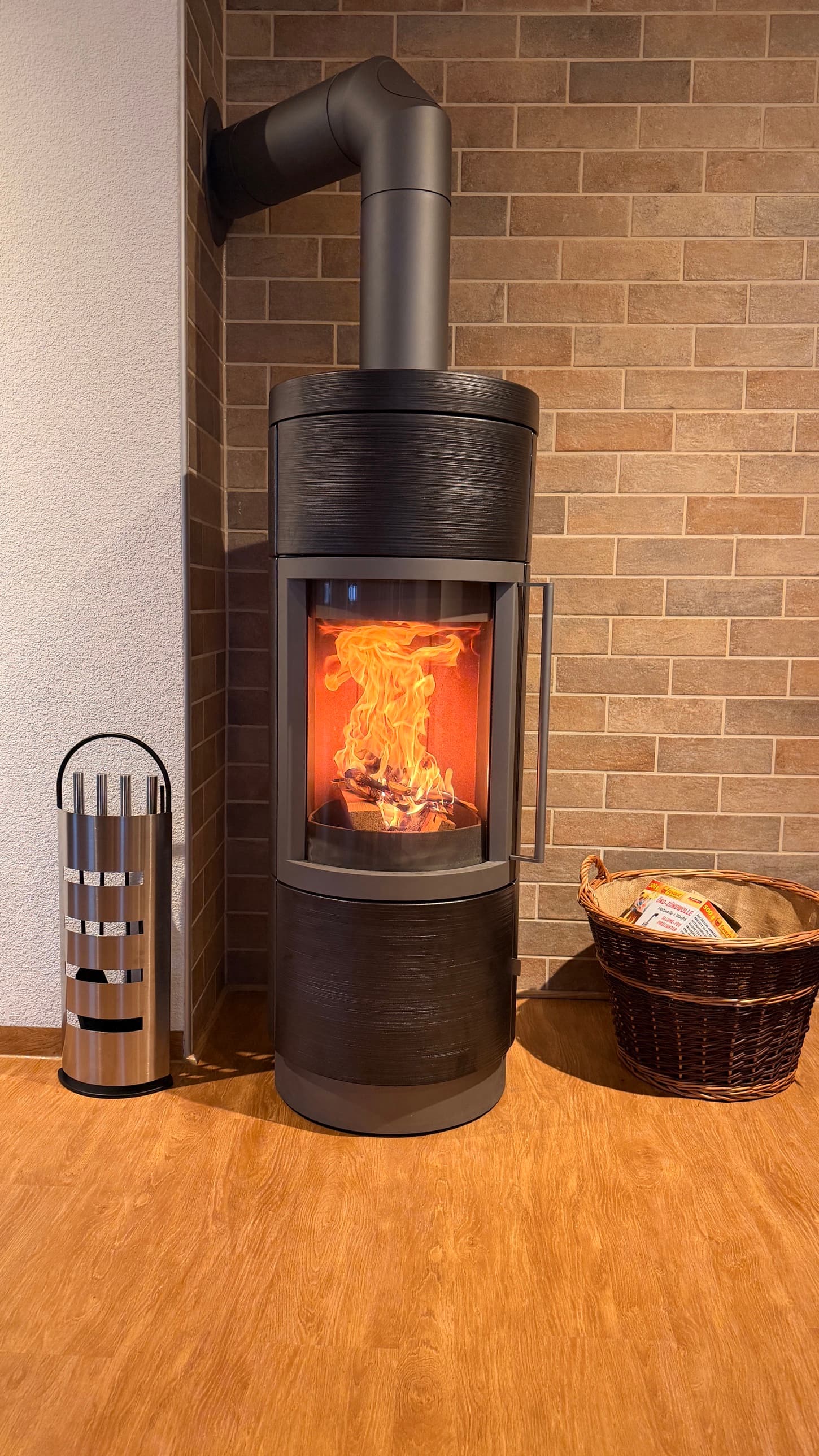 Hase Lima in einer Zimmerecke in Gerbrunn, runder Ofenkörper mit gebürsteter Keramik in Meteorit, Metallteile in Silbergrau, brennendes Feuer hinter der gebogenen Scheibe. Dahinter eine Wand in Ziegeloptik, links ein Kaminbesteck, rechts ein Weidenkorb.
