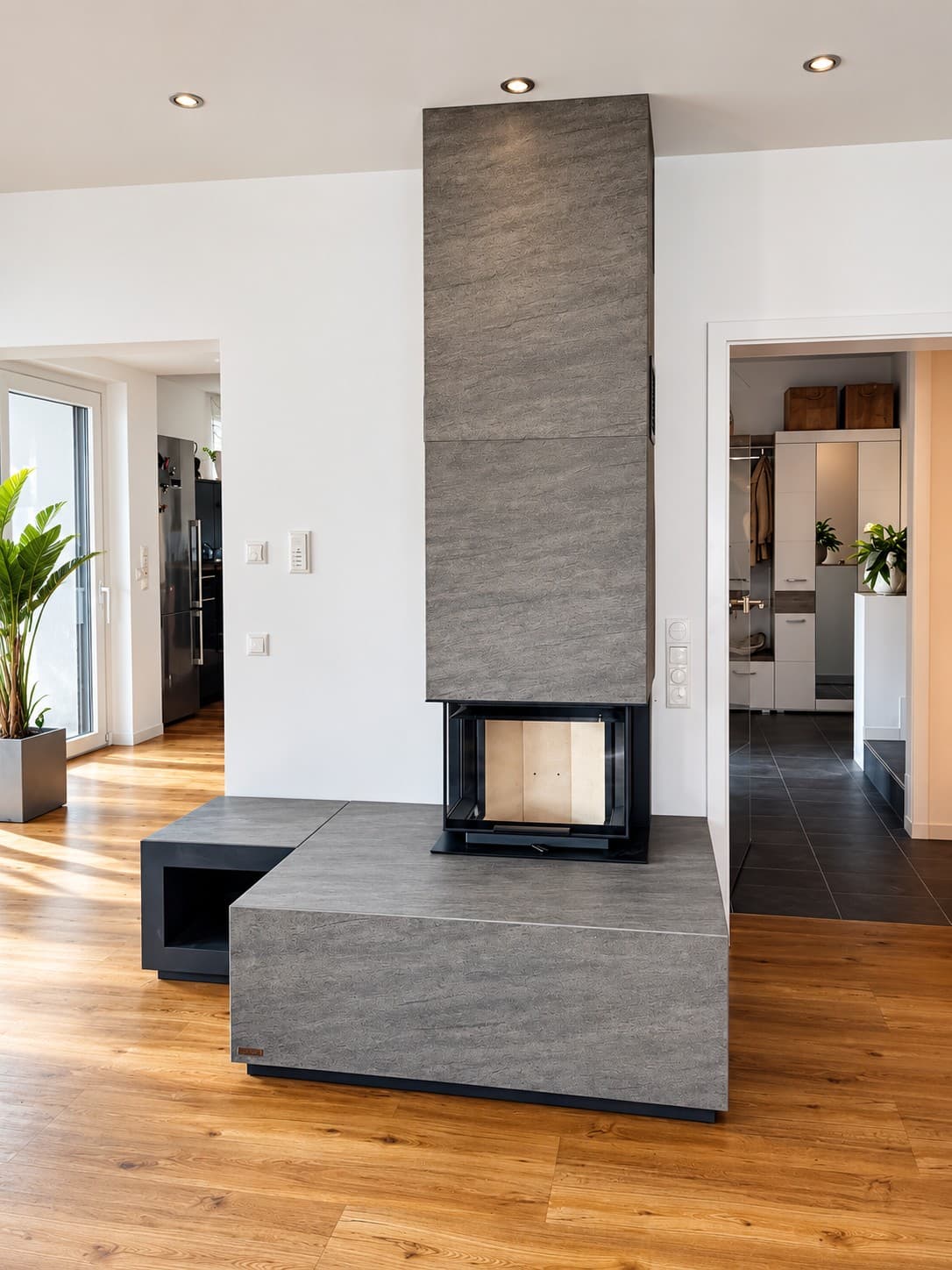 neocube P13 in Keramik Iron Grey, die Verkleidung läuft bis zur Decke, davor eine breite Ofenbank, links ein offenes Holzfach, im Hintergrund Küche und Flur.