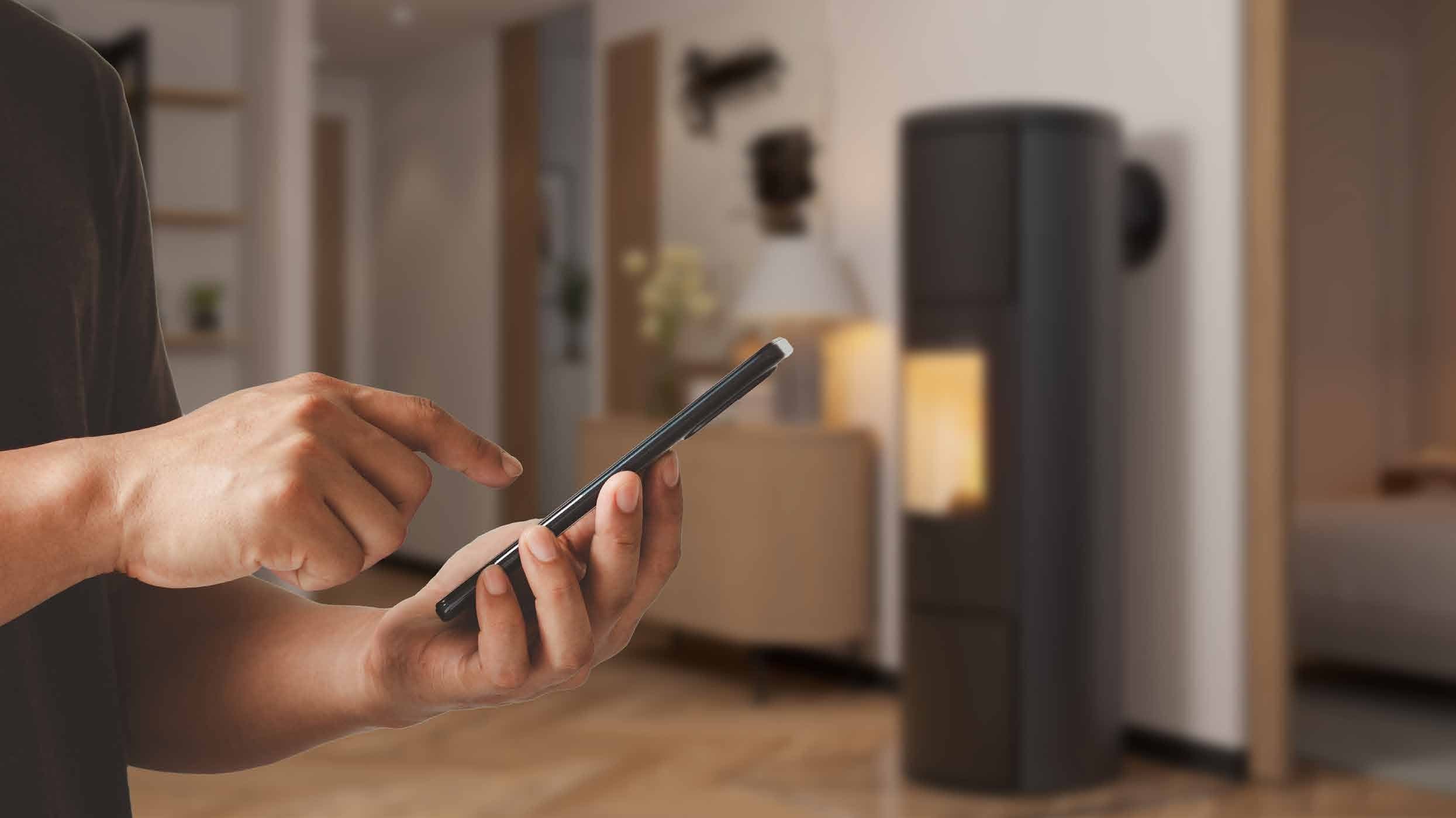 Person bedient die CB ABS Pro+ App am Smartphone, im Hintergrund ein brennender Kaminofen