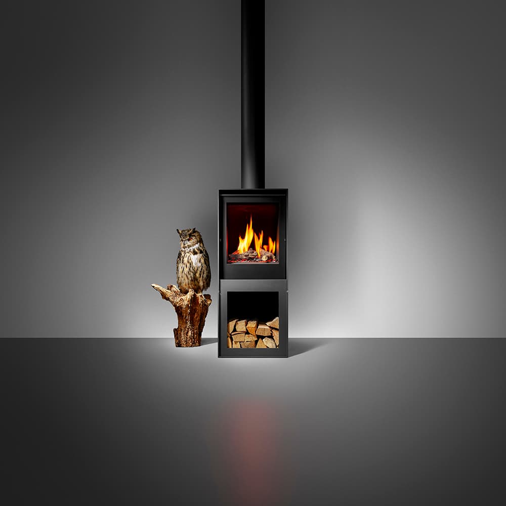 Kaminofen Barbas Box Gas 45 mit brennender Gasflamme, schwarzer Rahmen, Holzlager unten, neben einer goldenen Eulenskulptur