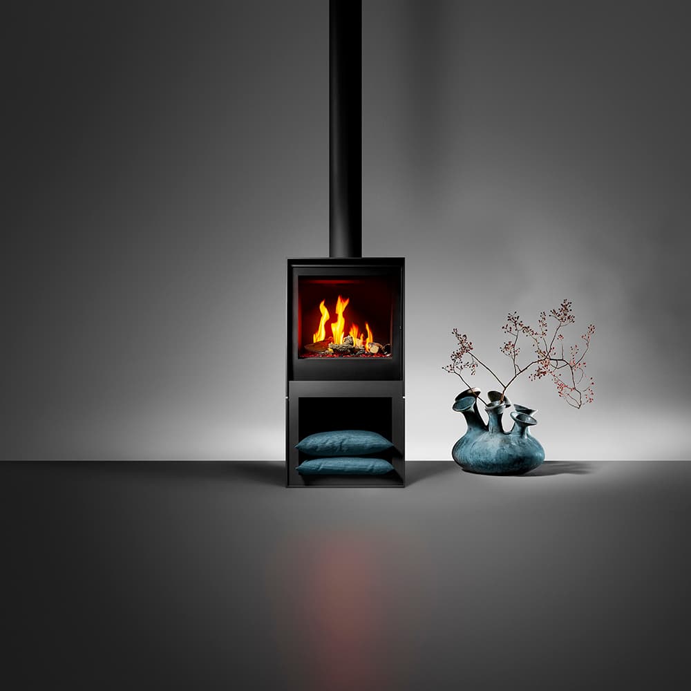 Barbas Box Gas 55 Gaskaminofen mit schwarzem Stahlgehäuse, loderndem Feuer und Abluftrohr, davor dekorative Kissen und Vase m