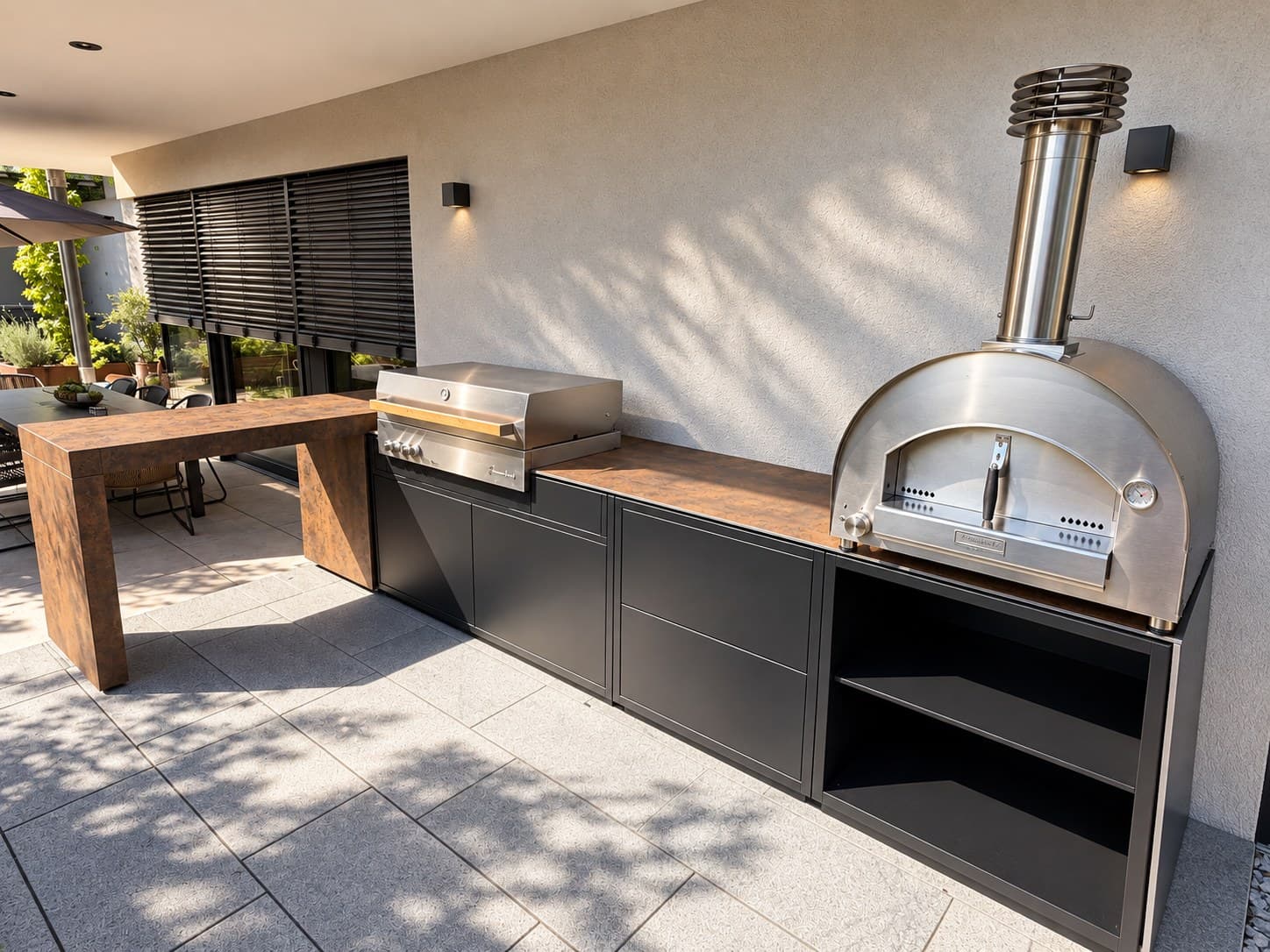 Modulare Outdoor-Küche von CB an einer Hauswand: schwarze Module mit Schubladen und offenem Fach, eingebauter Gasgrill Flammkraft Block D aus Edelstahl, rechts ein Pizzaofen aus Edelstahl mit Schornstein, links die Keramiktheke.