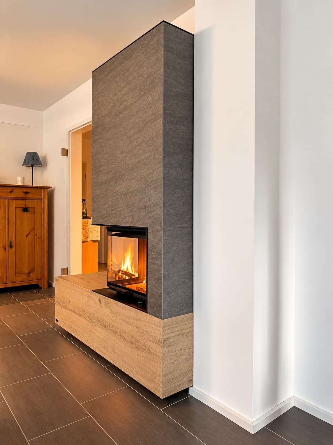 Seitenansicht des Eckkamins: Blick durch die seitliche Scheibe auf das Feuer, die Bank in Eichenoptik läuft nach vorn in den Raum.