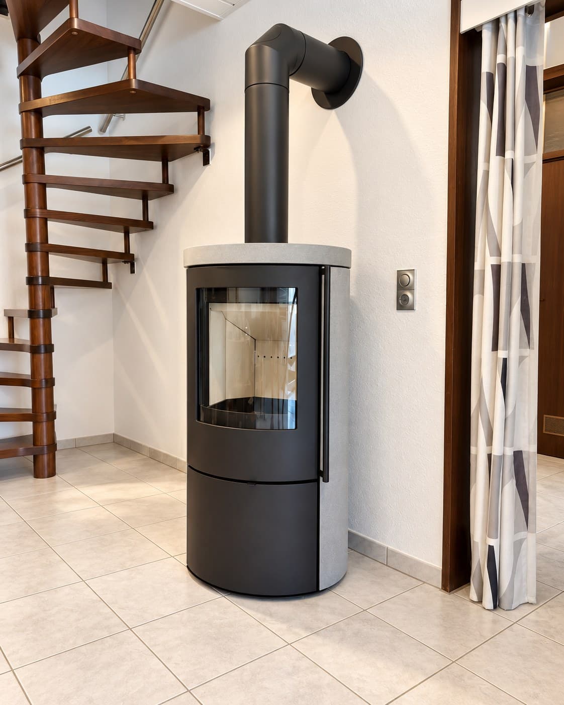 VAIO IQ Top 1 von CB-tec mit Natursteinverkleidung Grigio Augusta in einer Raumecke in Schweinfurt, daneben eine hölzerne Wendeltreppe, das Rauchrohr führt oben im Bogen in die Wand.