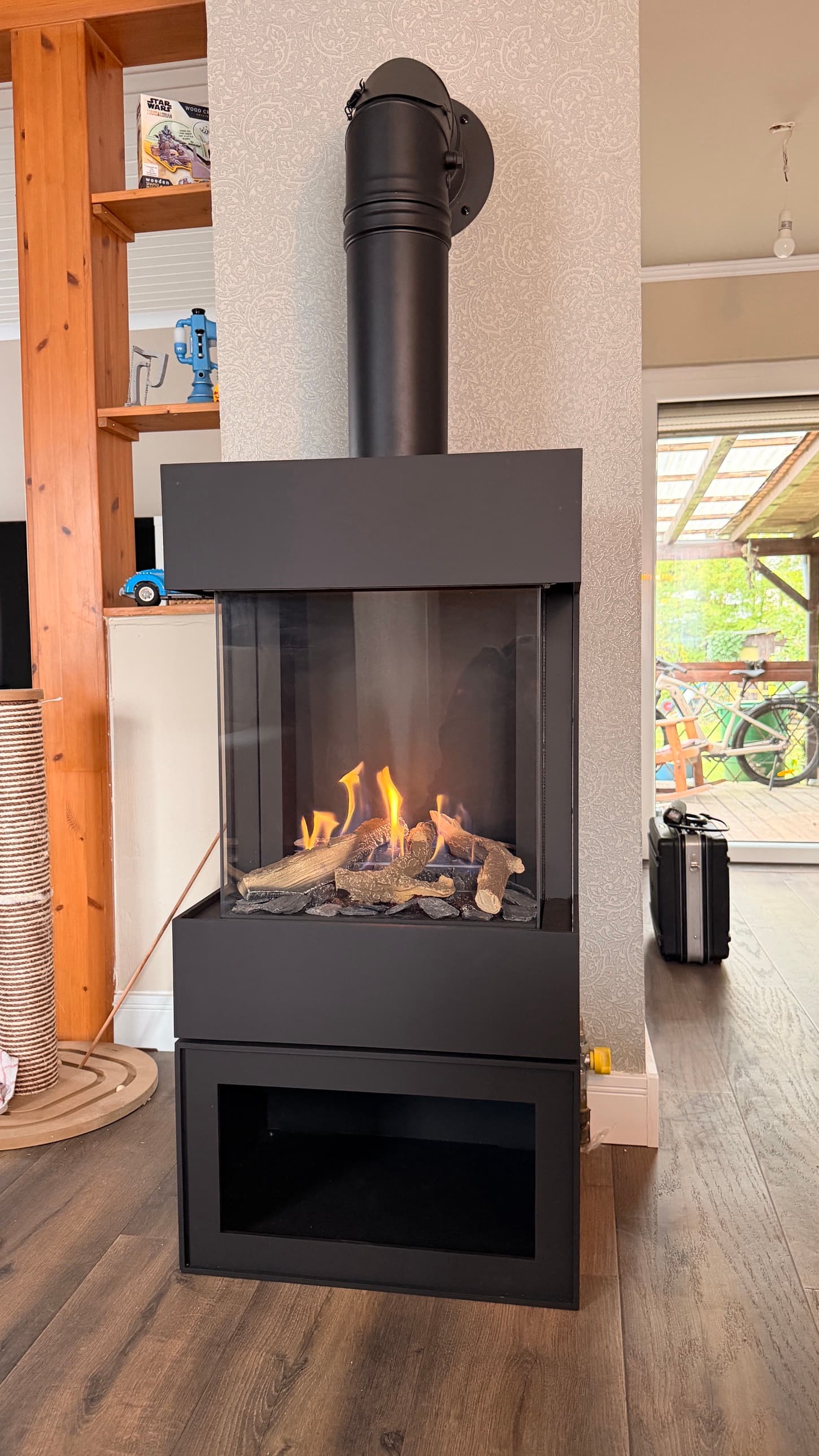 Barbas Box Gas Panorama 45/45 in schwarzem Stahl, dreiseitig verglast, mit brennendem Gasfeuer. Darunter ein Sockel mit offener Nische, darüber eine schwarze Haube, aus der das Rauchrohr senkrecht in die Wand führt.