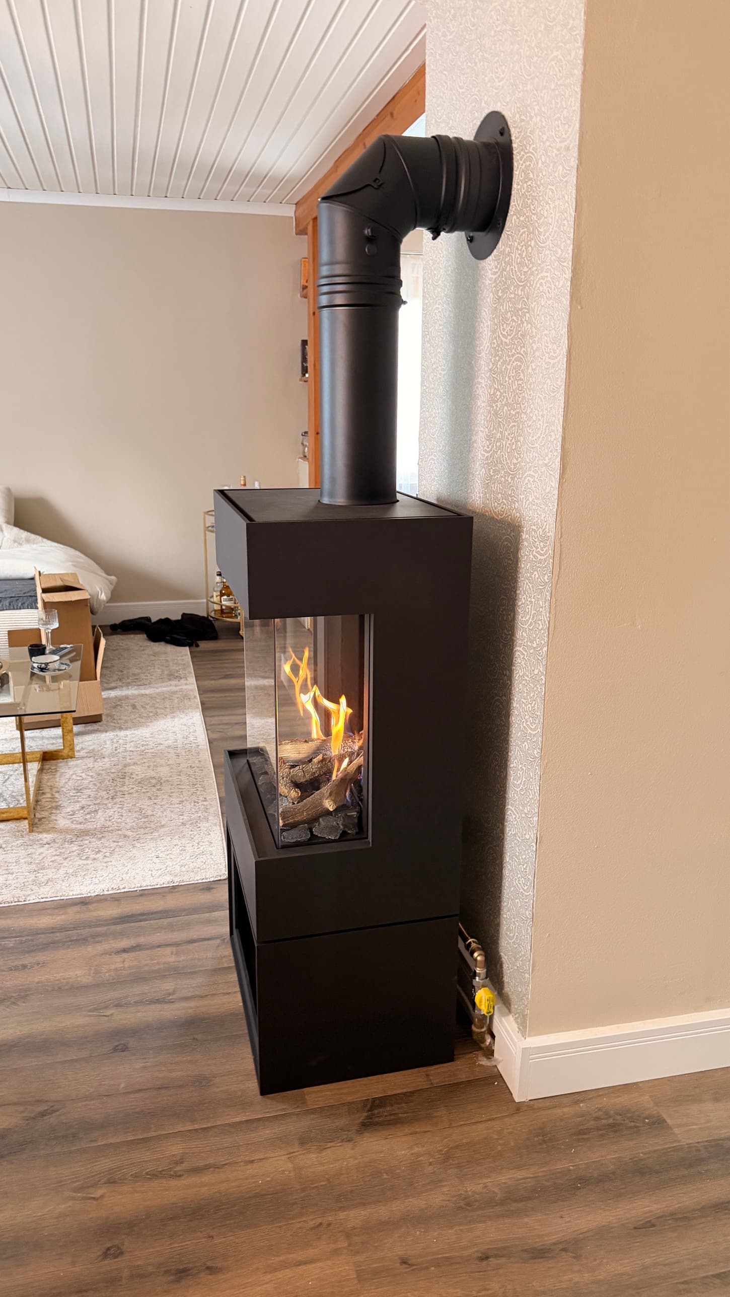 Derselbe Gaskamin aus einer anderen Richtung gesehen: Durch die seitliche Scheibe sind die Flammen zu sehen, das Rauchrohr führt im Bogen zur Wand.